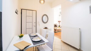 A modern kitchen with white cabinetry, a dining table with two chairs, a hanging light fixture, and a view into the living area. - Midtown Apartment by CorfuEscapes (Corfu)