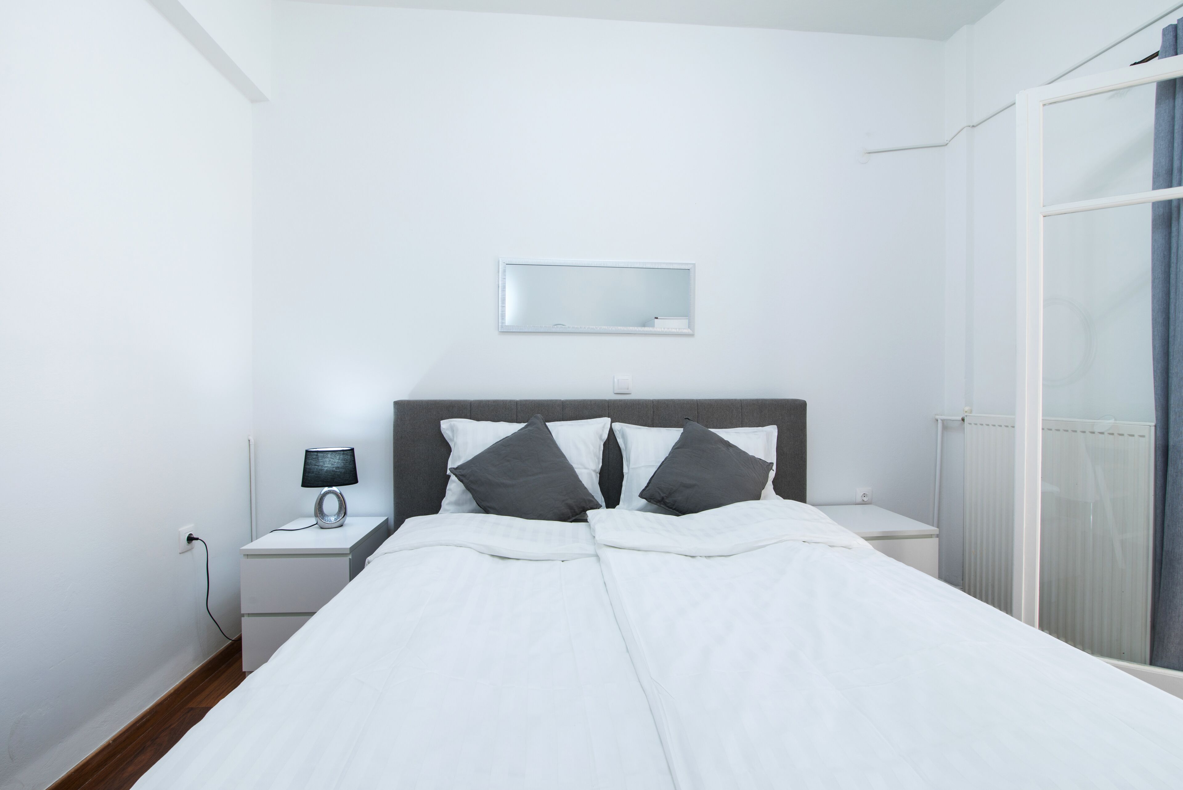 Un lit bien fait, avec une literie blanche et deux oreillers foncés, une table de chevet avec une lampe, un miroir fixé au mur et une fenêtre donnant sur un beau paysage.