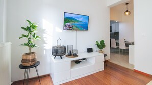 A modern living room with a flat-screen TV mounted on the wall, a white entertainment unit, a potted plant, and a dining area visible through an open doorway. - Midtown Apartment by CorfuEscapes (Corfu)