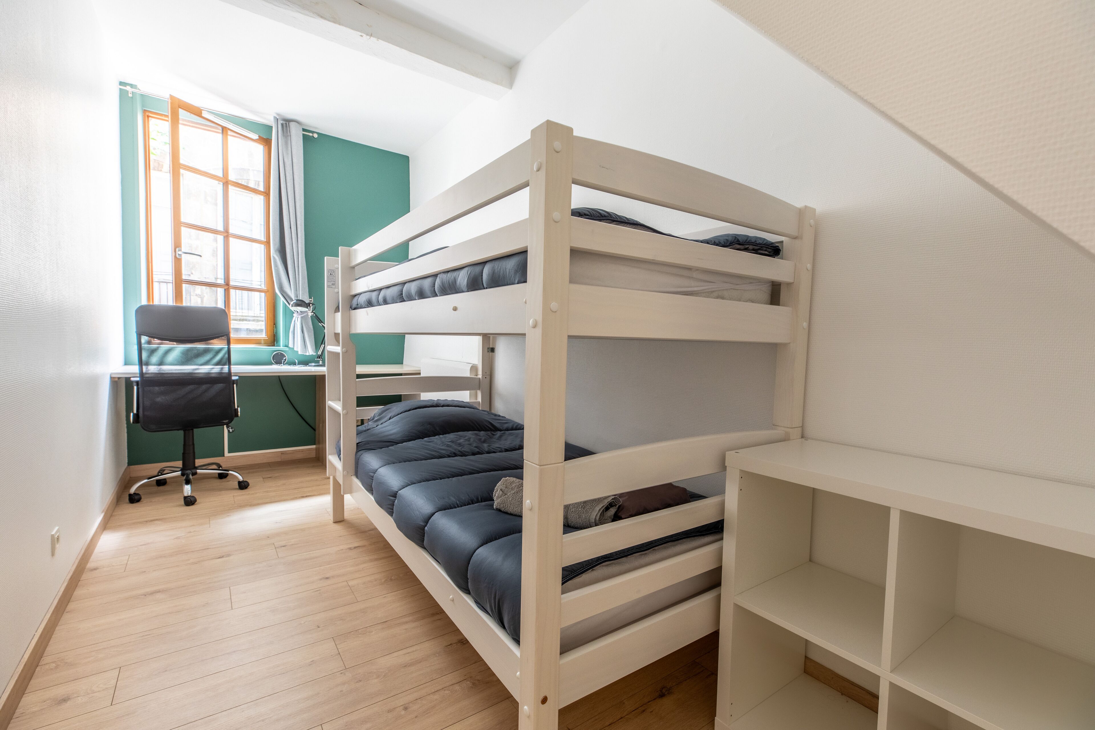 Een slaapkamer met stapelbed, bureau en stoel, een raam met gordijnen, en een licht houten vloer.