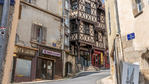Una calle estrecha con edificios tradicionales de entramado de madera, una señal de prohibido estacionar y una tienda cerrada con un letrero que dice "LICHANT DE LAGUOLE".