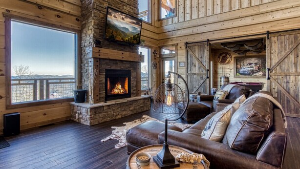 A spacious living room with a stone fireplace, leather sofa, and large windows offering a view of the mountains. - Moonshiners Hideout | Swing Bed, Hot Tub & Mountain Views (Gatlinburg)