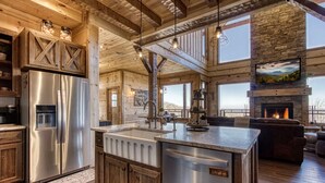A spacious kitchen with a large island, stainless steel appliances, and a stone fireplace. - Moonshiners Hideout | Swing Bed, Hot Tub & Mountain Views (Gatlinburg)