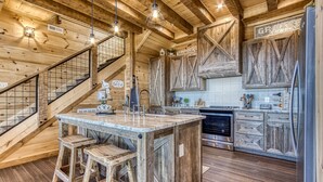 A rustic kitchen with wooden cabinetry, a central island with stools, and a staircase with a metal railing. - Moonshiners Hideout | Swing Bed, Hot Tub & Mountain Views (Gatlinburg)