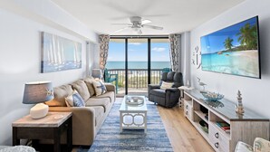A modern living room with a large TV, comfortable seating, a coffee table, and a view of the ocean.