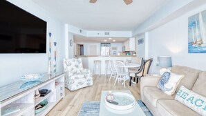A modern living room with a TV, a ceiling fan, a dining area with chairs, and a kitchen in the background.