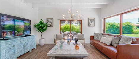 Uma sala de estar ampla, com uma TV de tela grande, uma mesa de centro, um sofá e um espaço de jantar com vista para o verde lá fora.