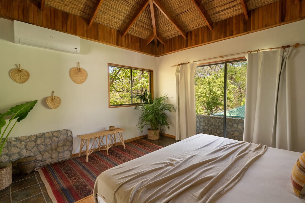 Een slaapkamer met een bed, een raam, een kamerplant, een bankje en een stenen muur.