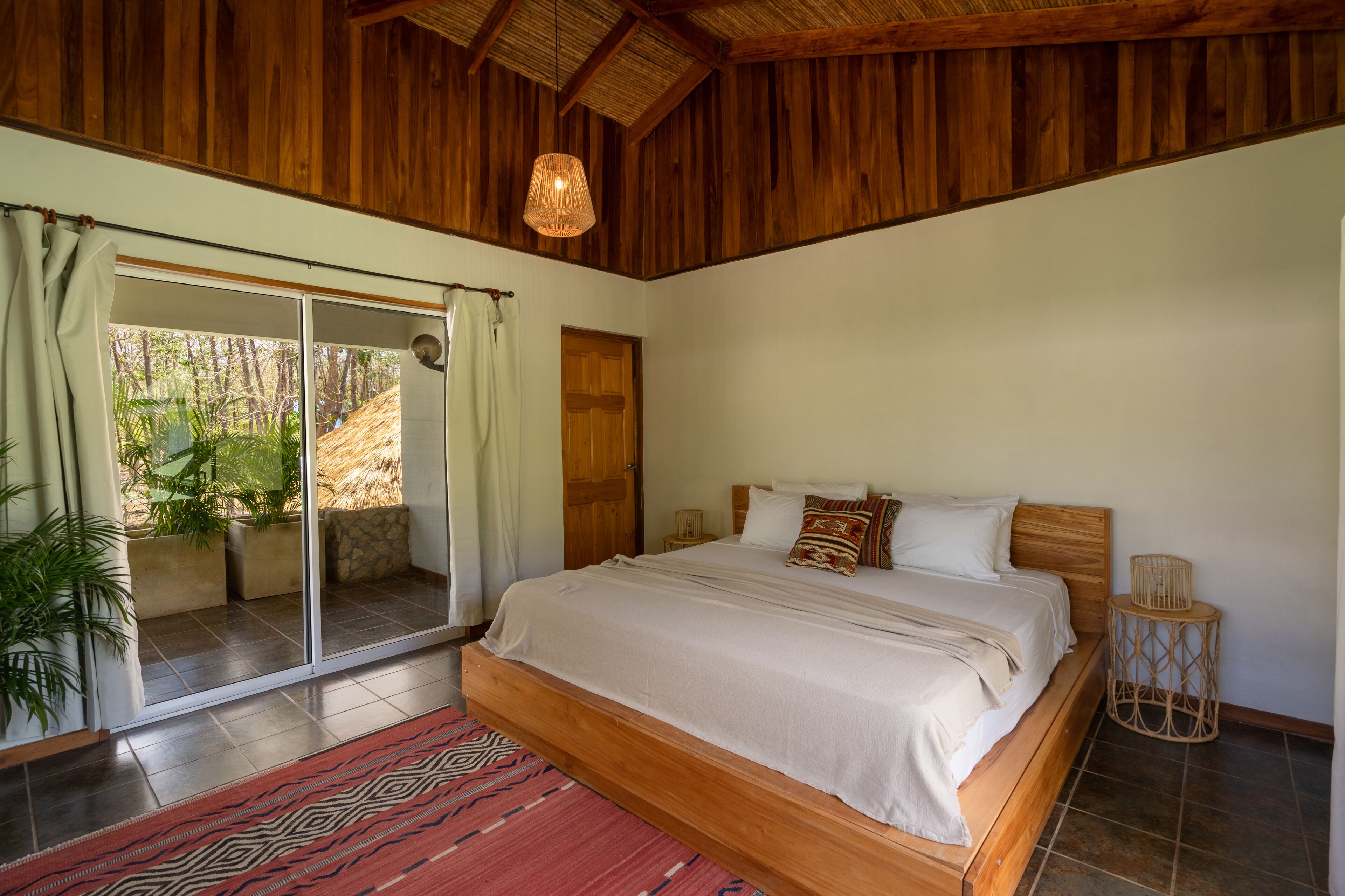 A spacious bedroom with a large bed, sliding glass doors leading to a balcony, and a wooden ceiling.