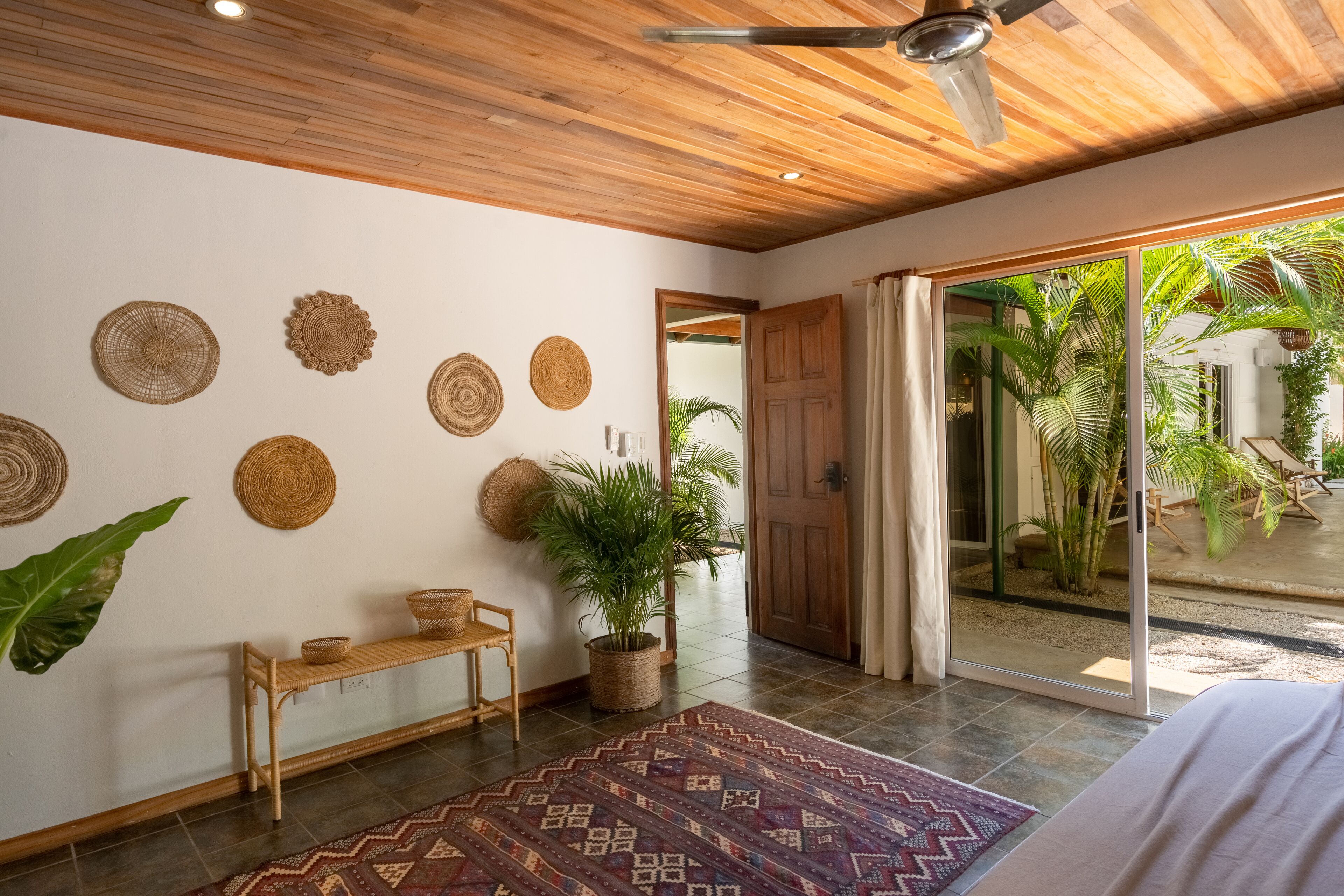 A room with a wooden ceiling, a bench, a patterned rug, and a door leading to a garden.