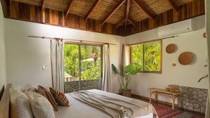 A bedroom with a large bed, two windows, a wooden ceiling, a stone wall, and a small table with a basket. - Dream Surf House Avellana (Tamarindo)
