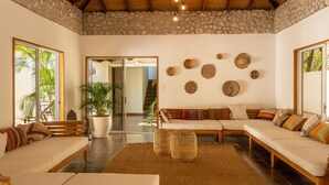 A spacious living area with a stone ceiling, wooden beams, and a large window. There are two sofas, a coffee table, and a potted plant. - Dream Surf House Avellana (Tamarindo)