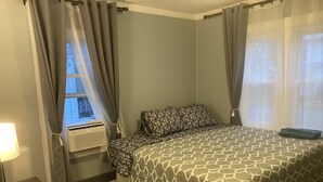 A bedroom with a bed, a window with curtains, an air conditioning unit, and a bedside table.
