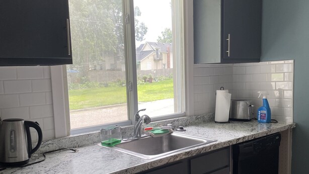 A kitchen with dark cabinets, a white countertop, a stainless steel sink, and a window with a view of a house and lawn.