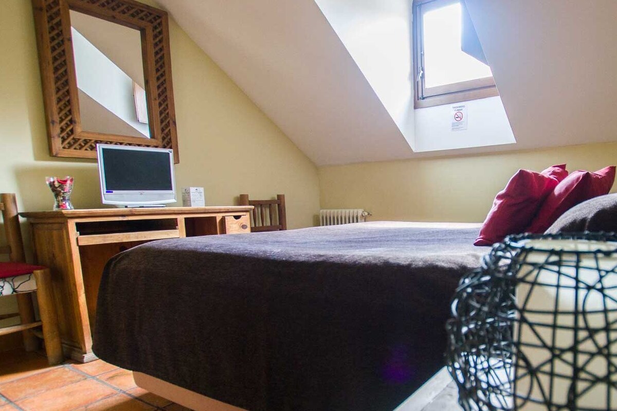 A bedroom with a sloped ceiling, a bed with dark bedding, a wooden desk with a laptop, a chair, a radiator, and a window with a view.