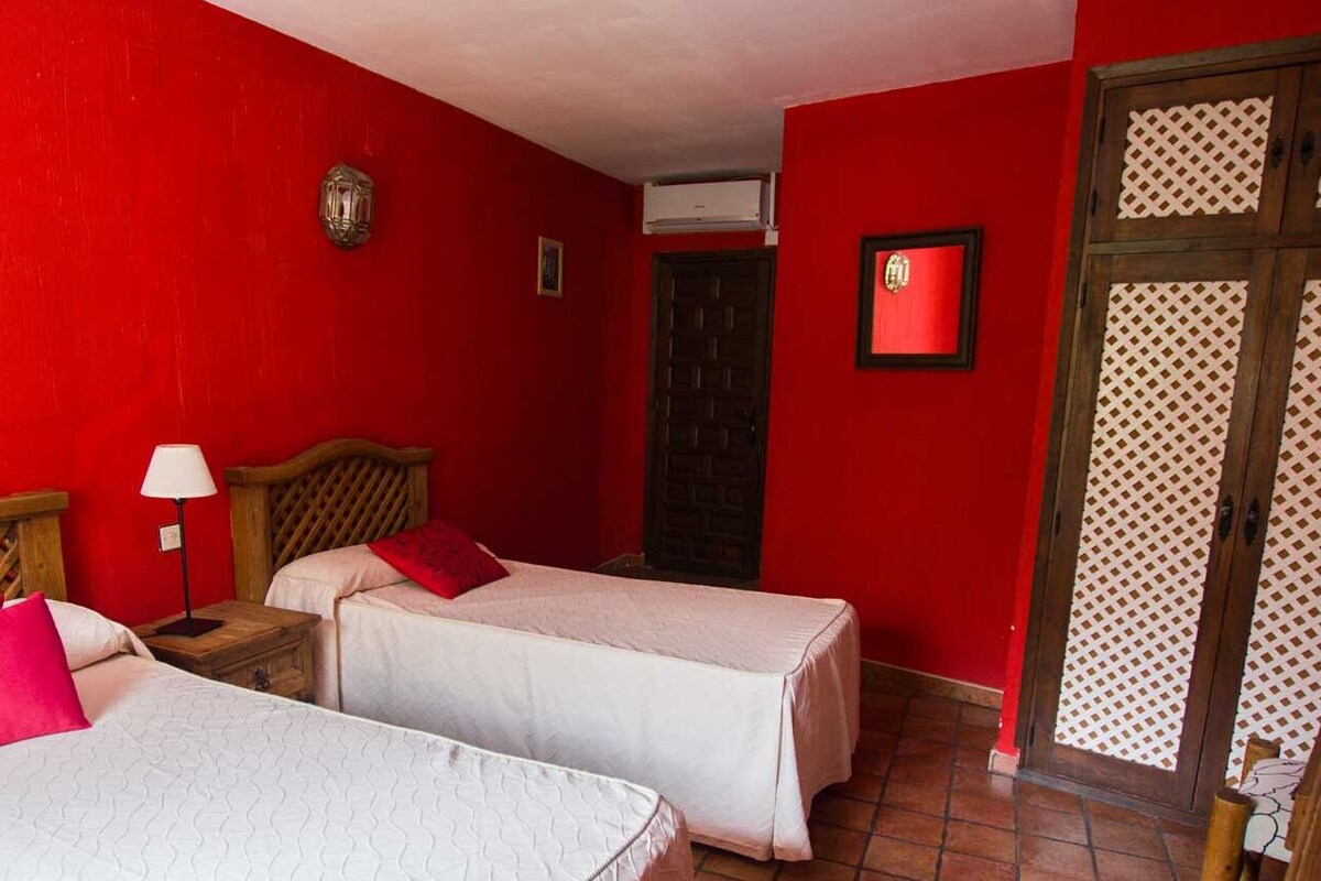 A room with two beds, a wooden headboard, a nightstand with a lamp, and a window with patterned curtains.