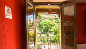 Una habitación con una pared roja, un ventanal con vistas a una colina y un balcón con barandilla de metal.