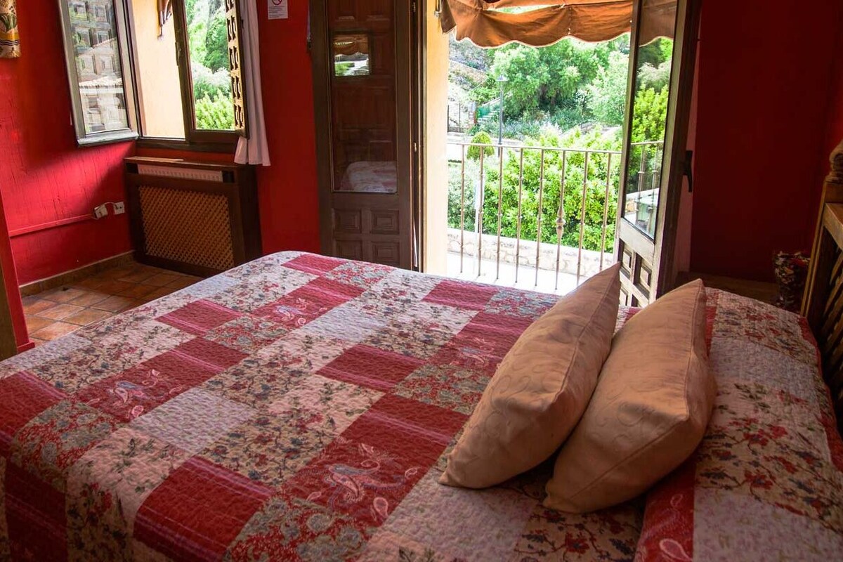 A bedroom with a red wall, a wooden bed, a quilt, and a view of greenery through a balcony door.