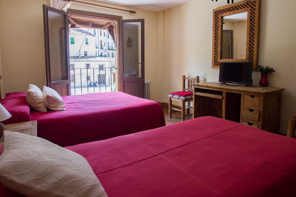 A hotel room with two beds, a desk, a chair, a TV, and a balcony with a view of buildings.