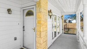 A covered porch with a white door, stone pillar, and two wall-mounted lanterns. - Charming, Hill Country Small-town Suite, Centrally located. (Harper)