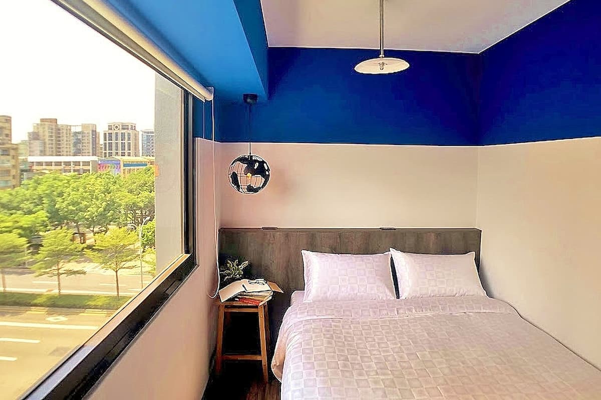 A bedroom with a large window, a bed, a bedside table, and a hanging light fixture.