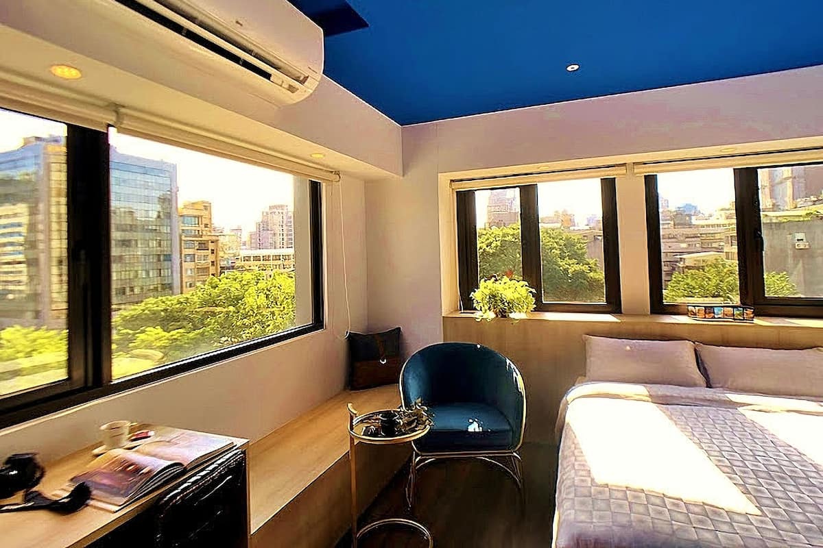 A modern bedroom with a bed, a desk, and a chair. There are large windows providing a view of the city.