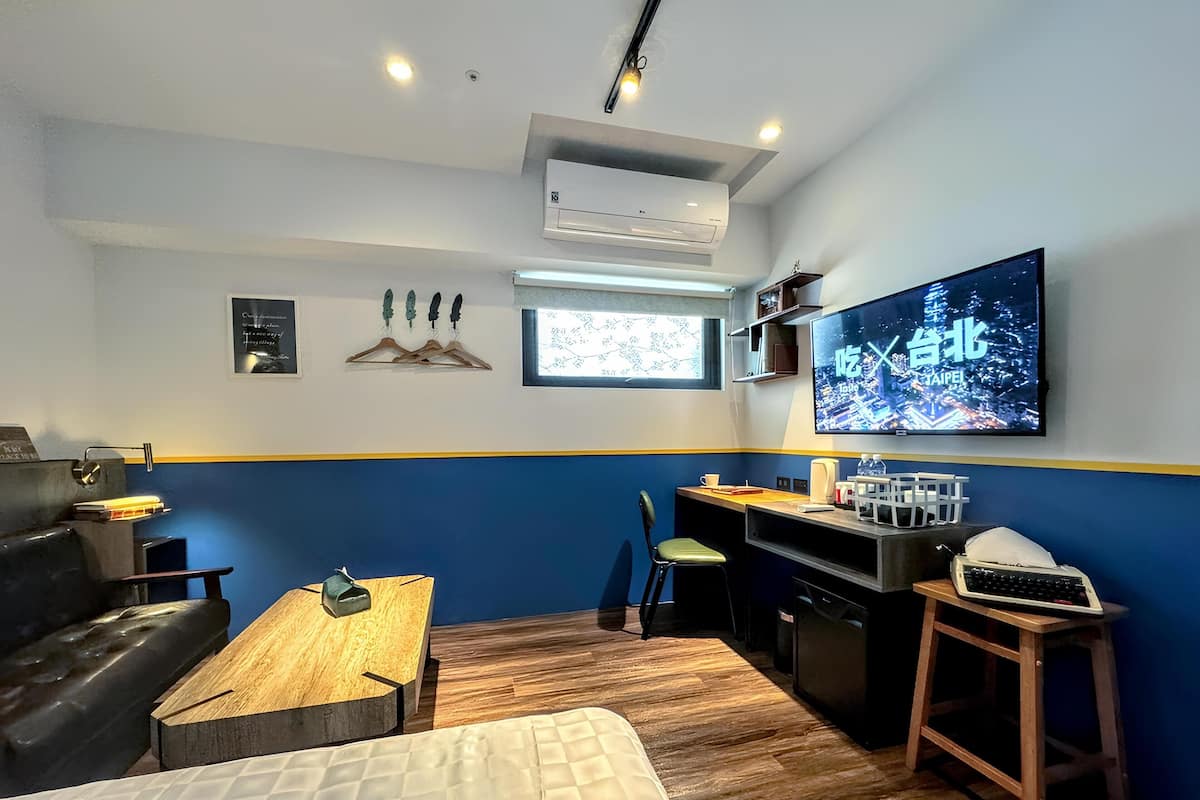A compact hotel room with a bed, a desk, a chair, a small kitchenette, and a TV displaying a cityscape.