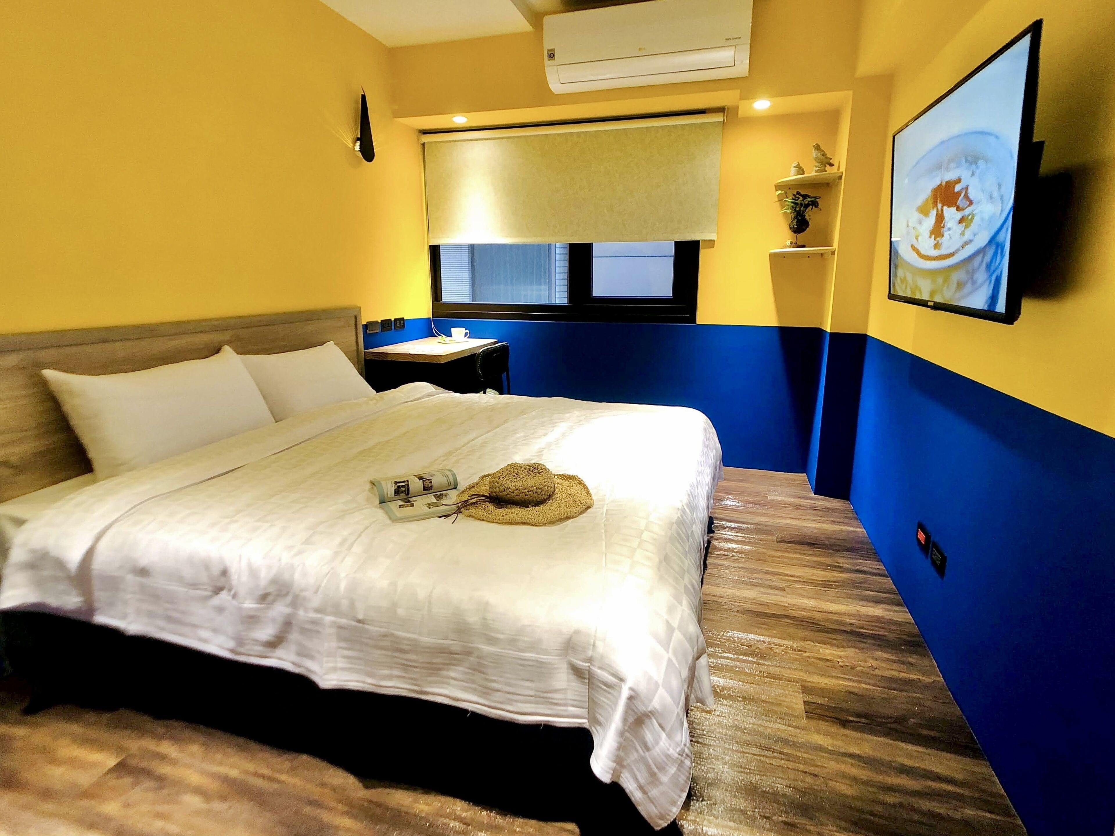 A hotel room with a bed, a nightstand, a window with blinds, a wall-mounted air conditioner, and a painting on the wall.