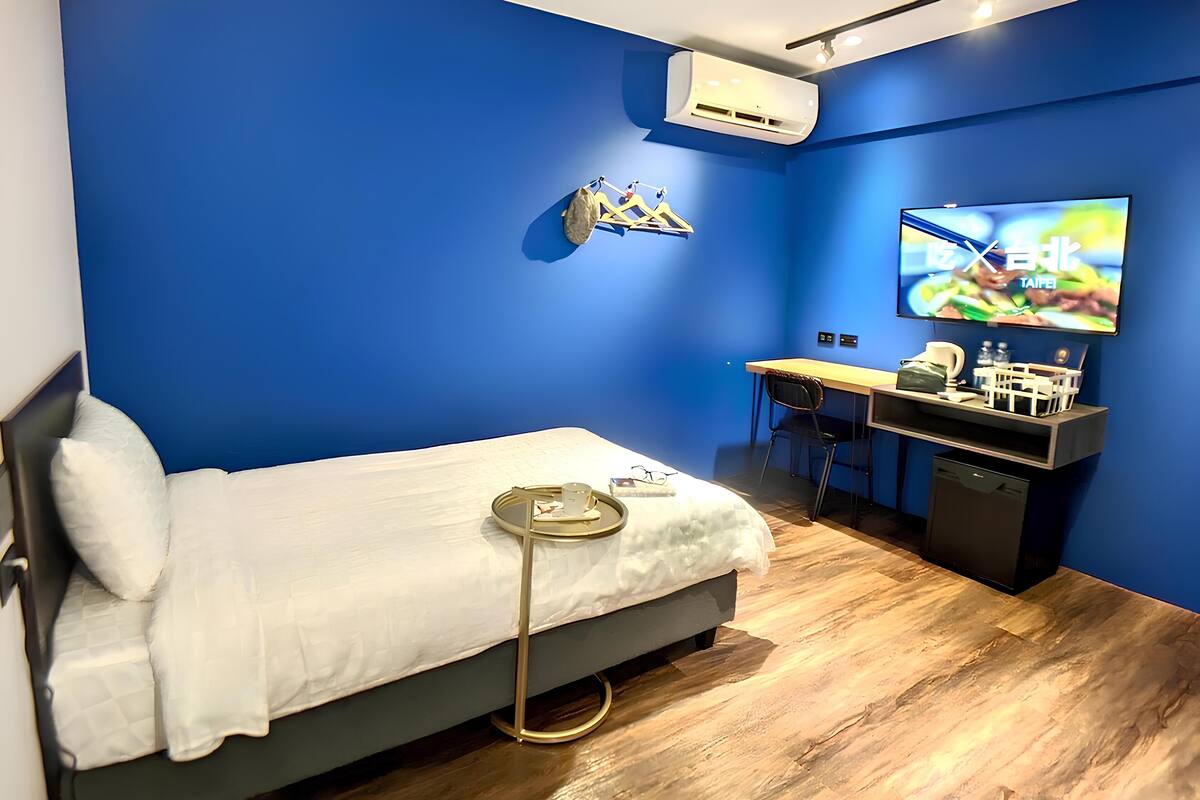 A hotel room with a bed, a desk, a chair, a TV, and an air conditioner.