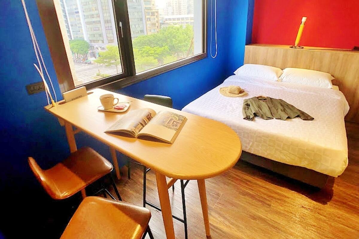 A room with a bed, a desk, and a chair. There is a window with a view of buildings and trees.