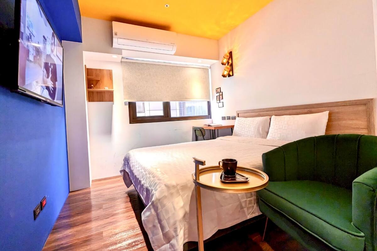 A hotel room with a bed, a green armchair, a small table with a cup, an air conditioning unit, and a TV mounted on the wall.