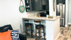 A kitchen with dark cabinets, a microwave, and a stainless steel refrigerator. There are two wooden bar stools and a small island with a granite countertop.