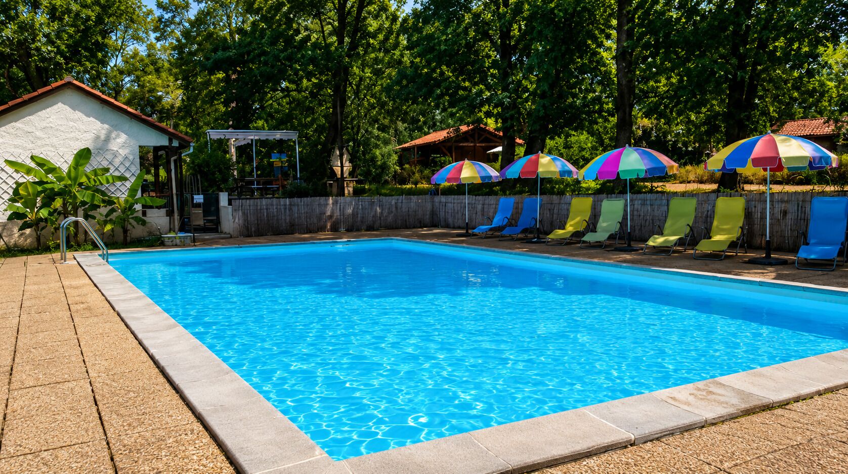Una piscina con borde de azulejos, rodeada por una valla de madera y protegida por sombrillas de colores.