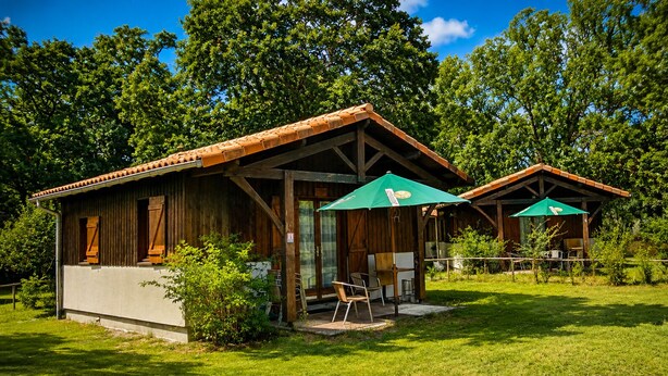 Two rustic wooden cabins with large windows and green umbrellas, surrounded by lush greenery. - Cottage surrounded by nature (Noaillan)