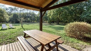 Une terrasse en bois avec une table de pique-nique et deux chaises longues, entourée d’une pelouse et d’arbres.