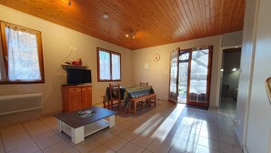 Un vaste salon doté d’un plafond en bois, d’un téléviseur posé sur un meuble en bois, d’une table à manger avec des chaises et d’une porte-fenêtre coulissante donnant sur l’extérieur.