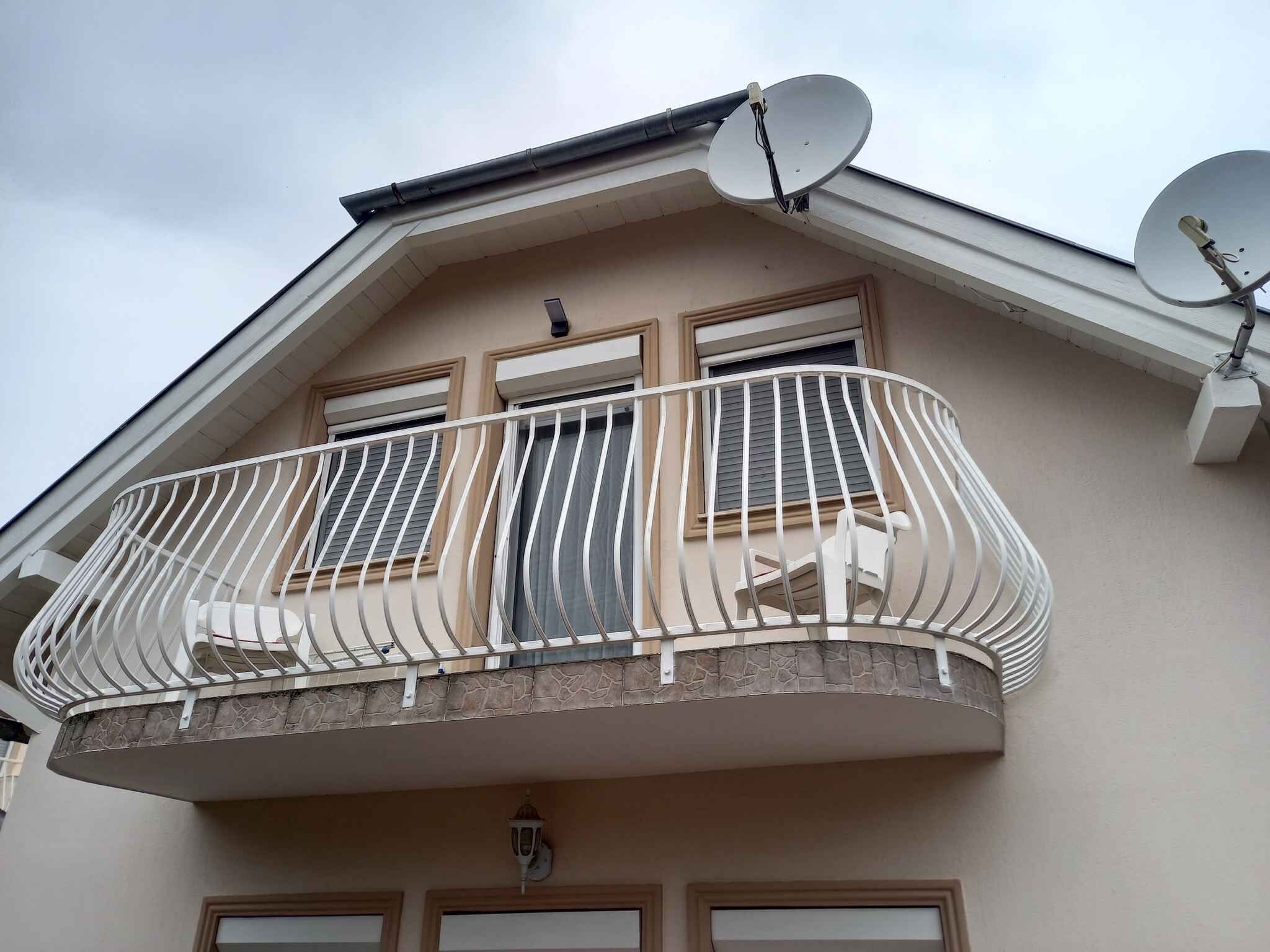 Un edificio residencial con balcón, barandilla metálica blanca, dos antenas parabólicas y un muro de piedra decorativo.