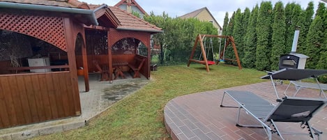 A backyard with a wooden pergola, a swing set, a barbecue grill, and a brick patio.