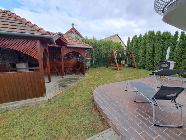A backyard with a wooden pergola, a swing set, a barbecue grill, and a brick patio.