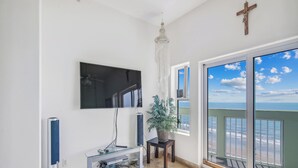 A living room with a TV, a chandelier, a plant, and a balcony with a view of the ocean. - Ocean Grove 704-Ocean View by Ryson Vacation Rentals (Galveston)
