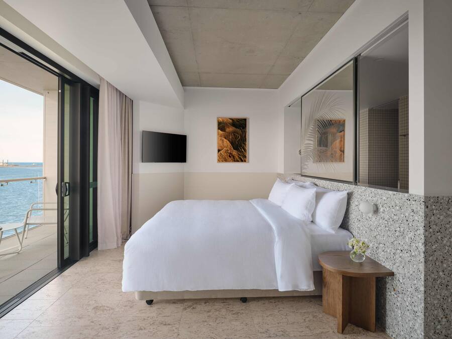 A modern hotel room with a large bed, a TV mounted on the wall, a small table with a vase, and a view of the sea through a sliding door.