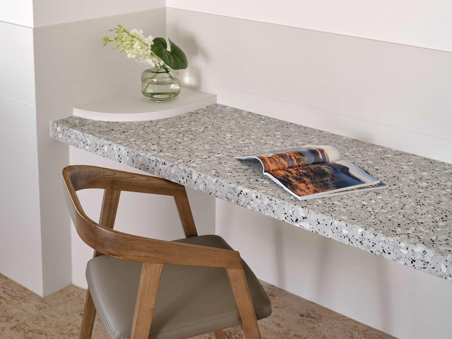 A corner desk with a speckled countertop, a wooden chair, and a vase with flowers.
