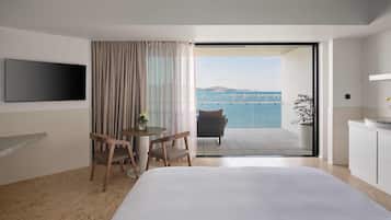 A hotel room with a bed, a TV, a table with chairs, a potted plant, and a view of the sea with boats.