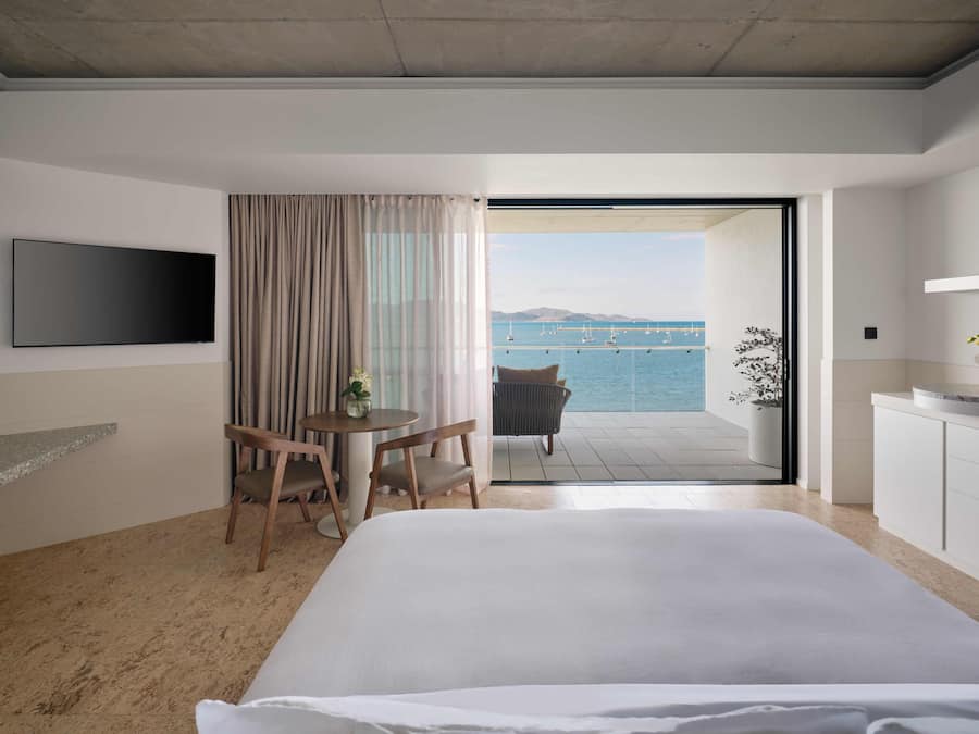 A hotel room with a bed, a TV, a table with chairs, a potted plant, and a view of the sea with boats.