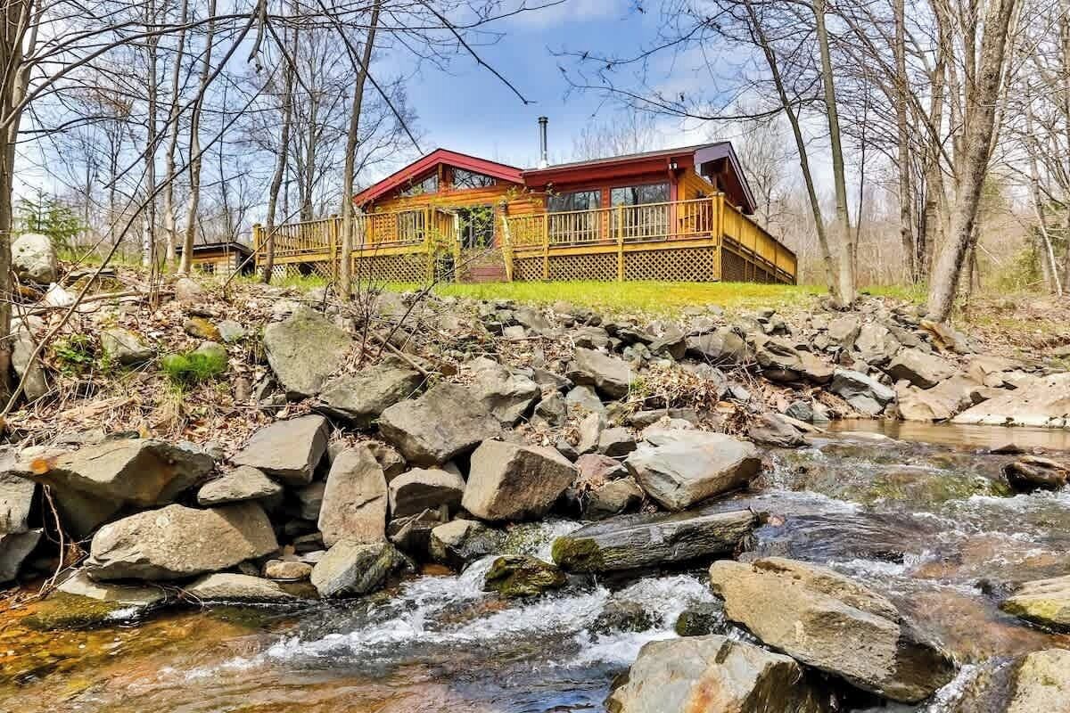 A cabin with a wooden deck and a red roof, surrounded by a rocky stream and trees.