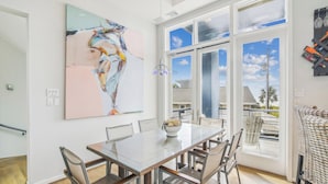 Un área de comedor con una mesa y sillas de vidrio, un cuadro abstracto grande en la pared, y vistas al exterior a través de amplias ventanas.