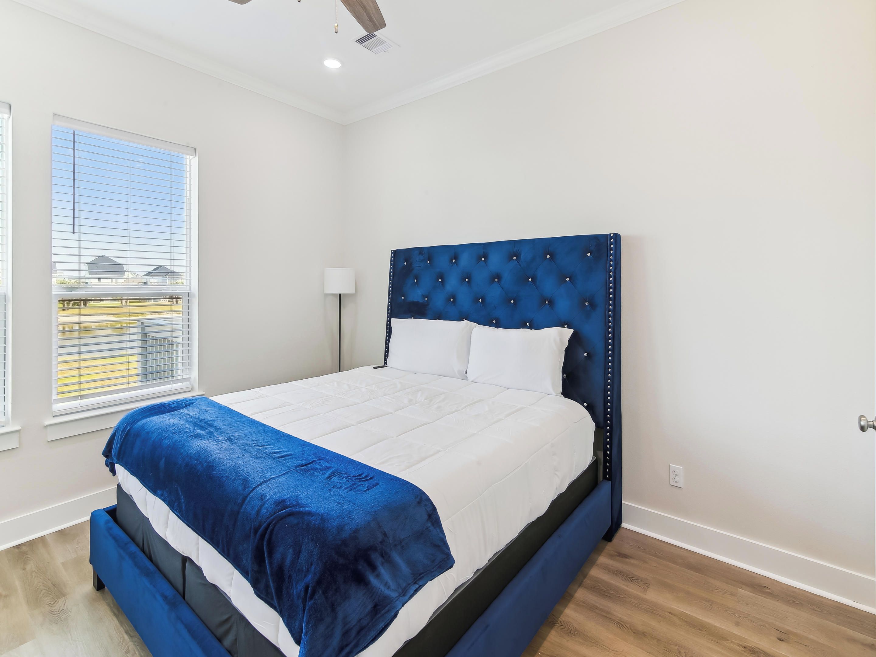 Une chambre à coucher avec un lit, une fenêtre avec des stores, une lampe sur pied et un ventilateur de plafond.