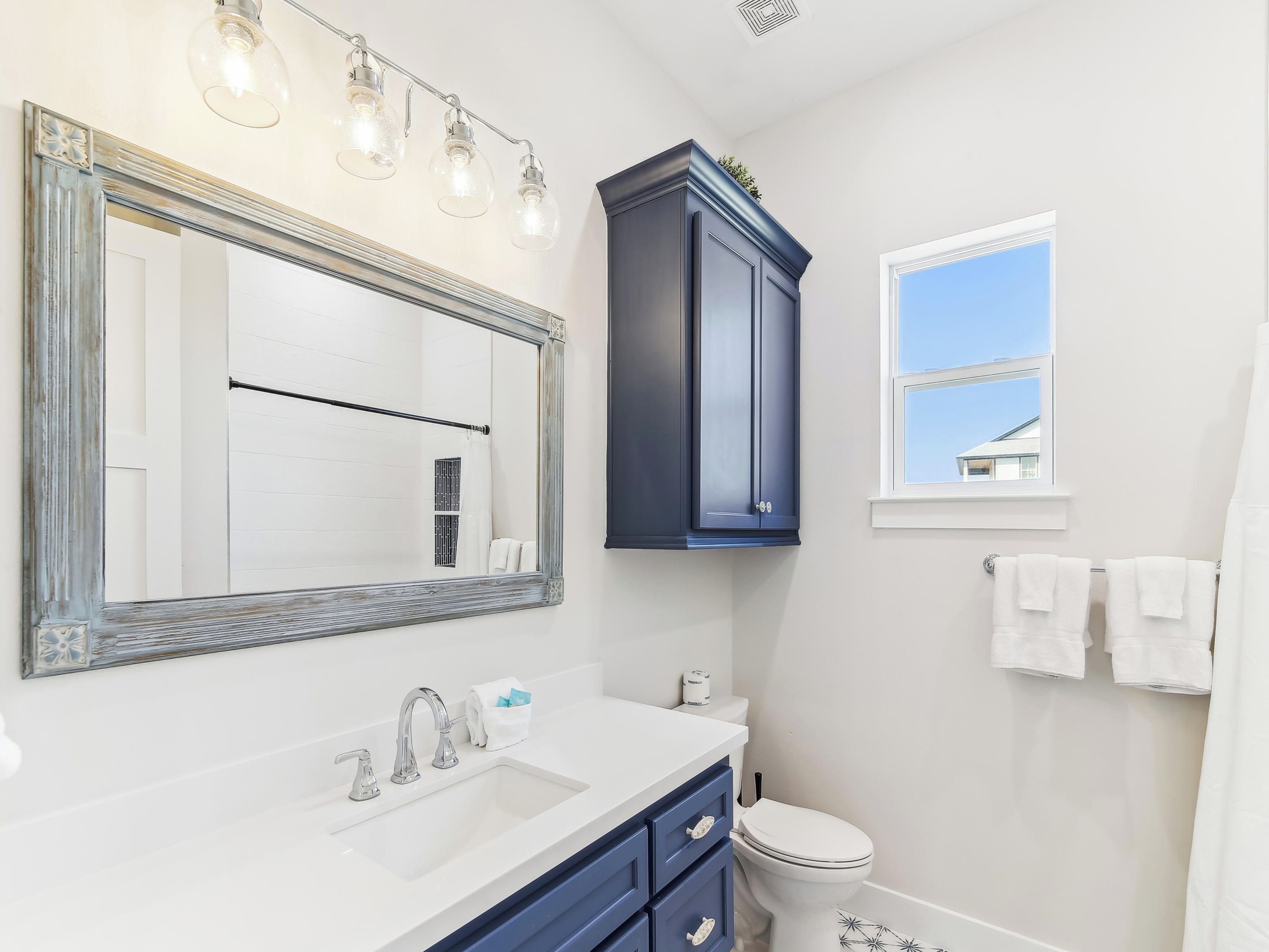 A bathroom with a white sink, two faucets, a large mirror with light fixtures, a dark blue cabinet, a toilet, and a window with a view of a house.