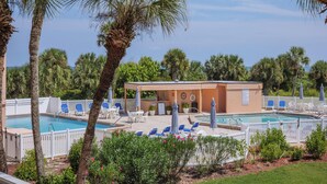 Zona de piscina con sillones de descanso y sombrillas, rodeada de palmeras y un edificio con un espacio de estar cubierto.