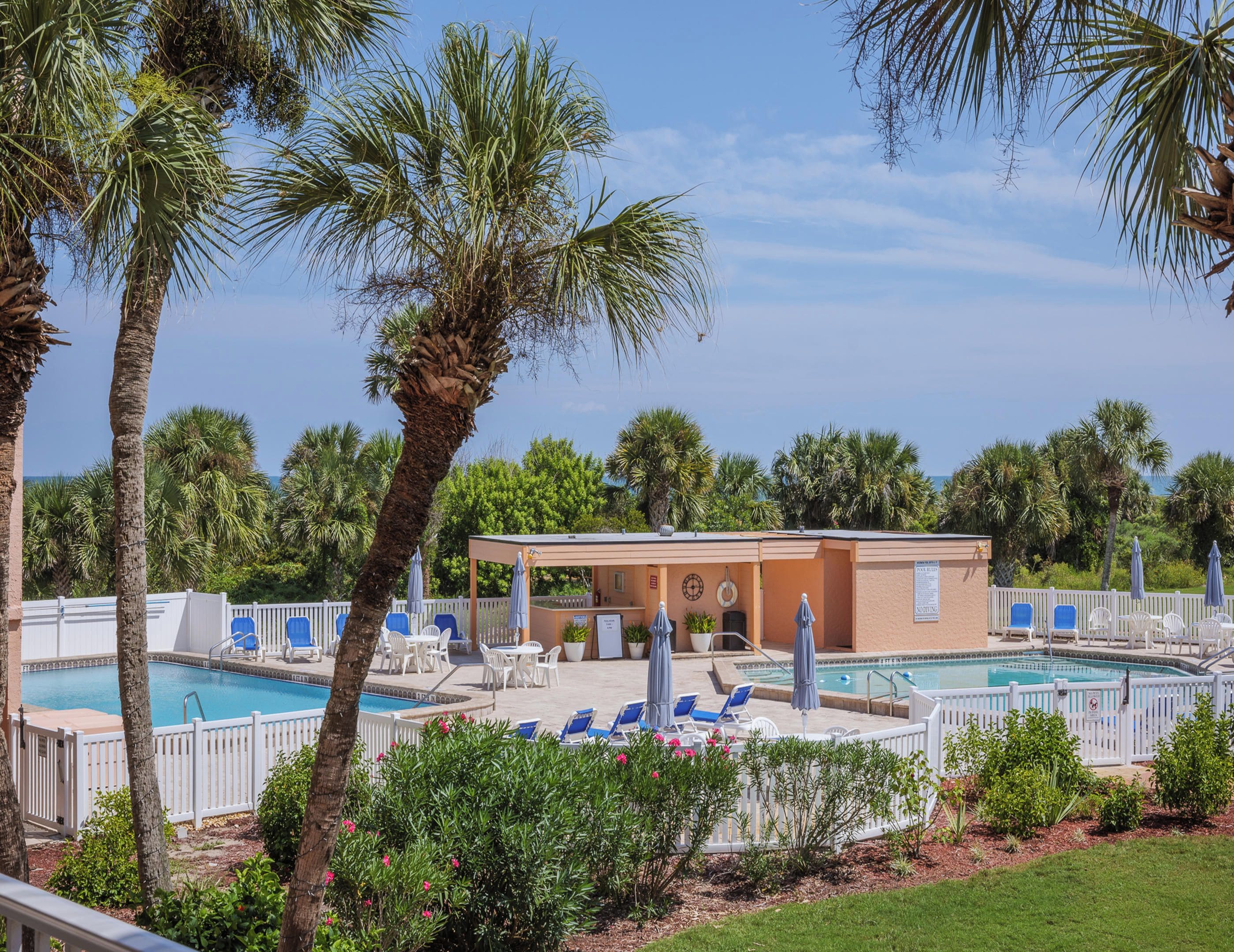 A pool area with lounge chairs and umbrellas, surrounded by palm trees and a building with a covered seating area.